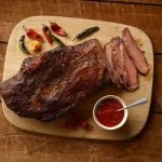 A top view of SADLER'S SMOKEHOUSE® whole brisket on a cutting board with some chilies and a ramekin or bbq sauce.