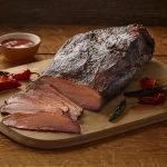 A side view of SADLER'S SMOKEHOUSE® whole brisket on a cutting board with some chilies and a ramekin or bbq sauce in the background.