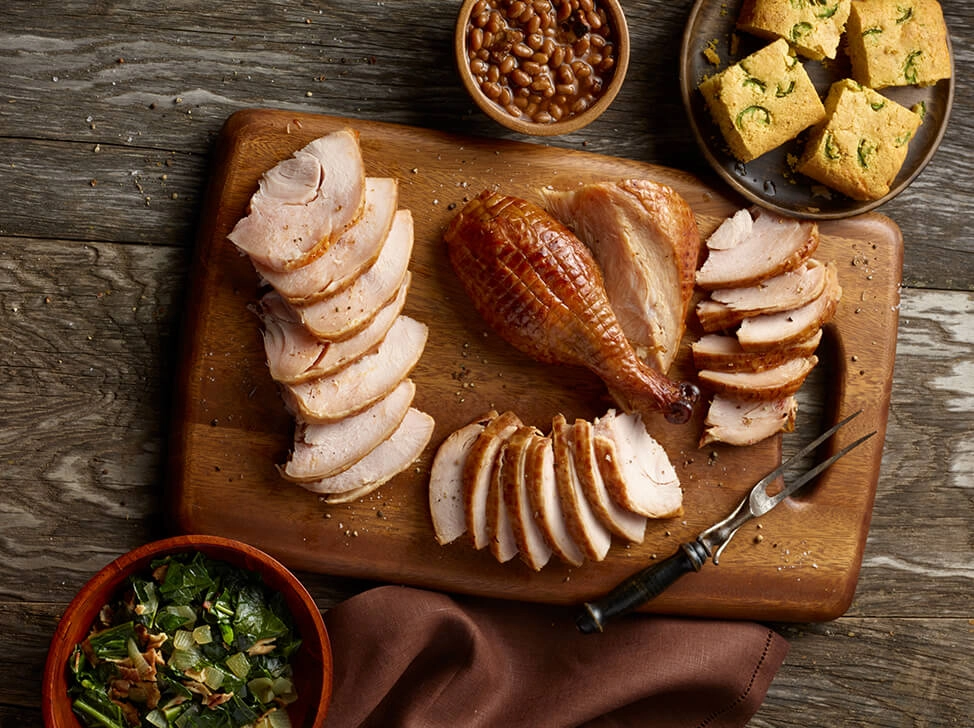 A carved SADLER'S SMOKEHOUSE® whole turkey on a cutting board with sides of corn bread, salad, and baked beans in the background.